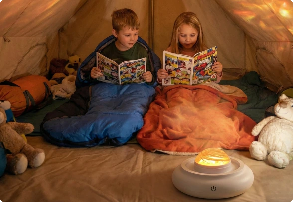 Two children sitting inside a tent, reading books while wrapped in blue and orange sleeping bags, with a glowing white projector lamp casting light on the tent floor. Stuffed animals are scattered around them.