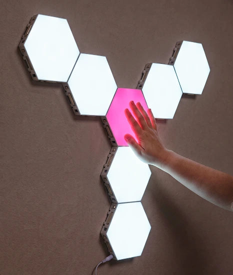 A hexagonal wall light panel set mounted on a brown wall, with one central panel glowing pink and a person's hand touching it. The surrounding panels are illuminated white.