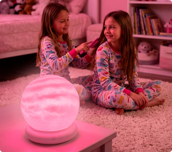 Two girls in matching unicorn-print pajamas sit on a pink rug in a bedroom, smiling while one brushes the other's hair. A glowing pink moon lamp sits on a small table in the foreground.