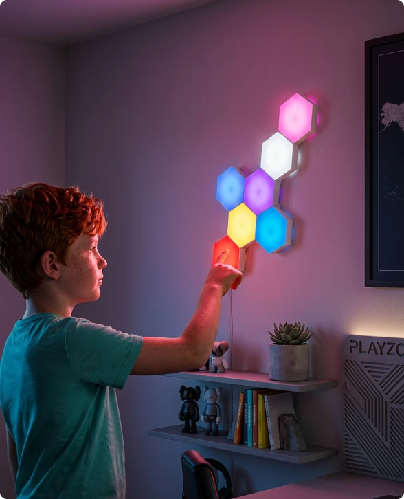A child in a teal shirt adjusts a colorful wall light set made of hexagonal panels mounted on a purple wall in a modern room.